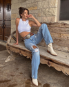 Woman posing in casual style with jeans and a white tank top on a wooden bench.