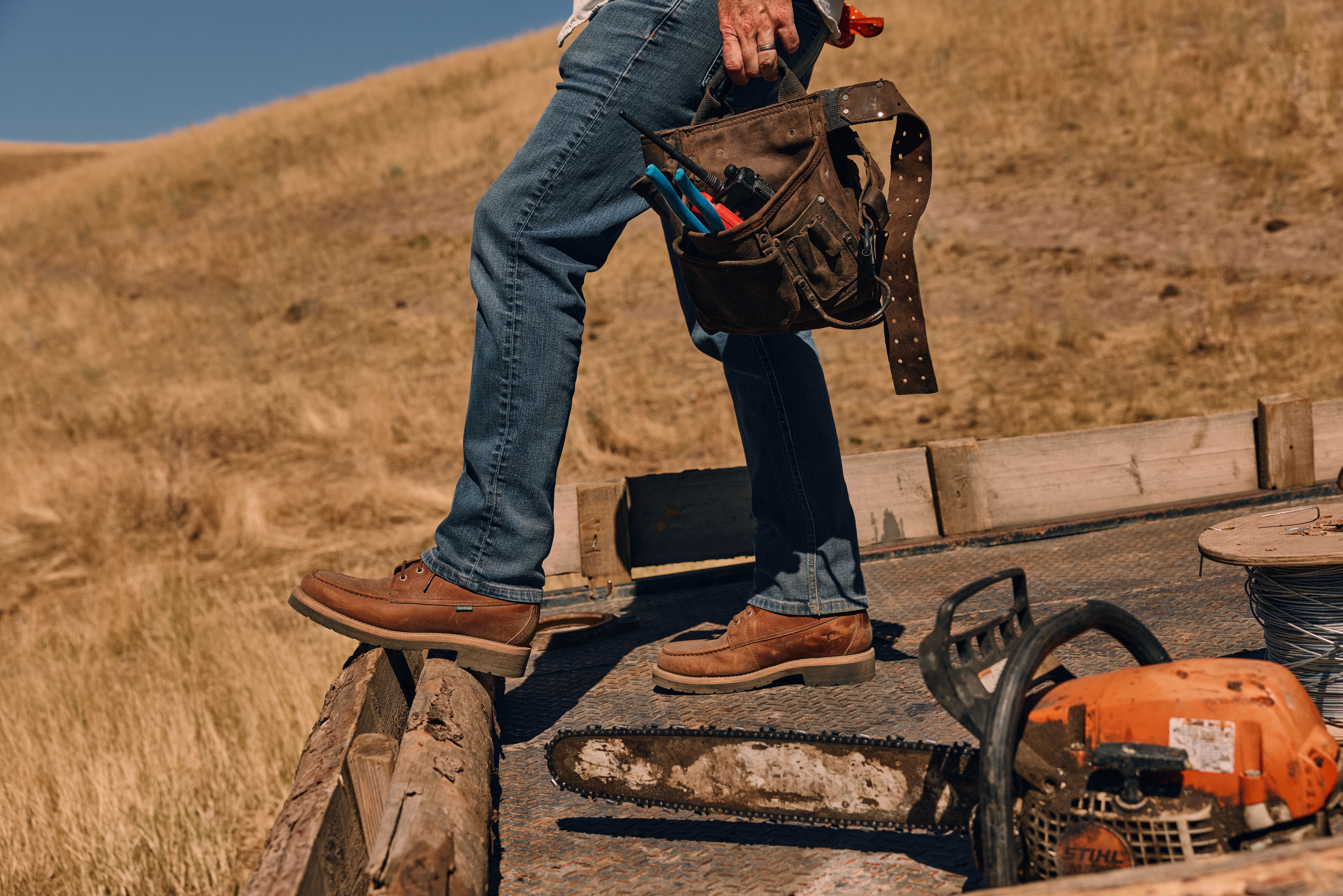 Person in jeans and work boots standing on a flatbed trailer, holding a tool belt, with a chainsaw and spool of wire beside them in a dry, grassy landscape.