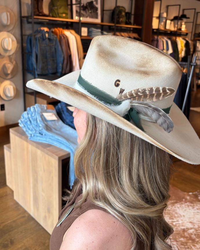 A woman with wavy blonde hair wears a light-colored cowboy hat adorned with a feather and initials "GW" in a clothing store.