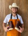 A man in a white shirt and cowboy hat stands against a wooden background, holding a pair of brown leather boots with light-colored soles. He wears an orange apron and glasses and is smiling.