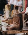 A display of various cowboy boots on a wooden table at an outdoor market, with some boots placed on boxes and hats visible in the background.