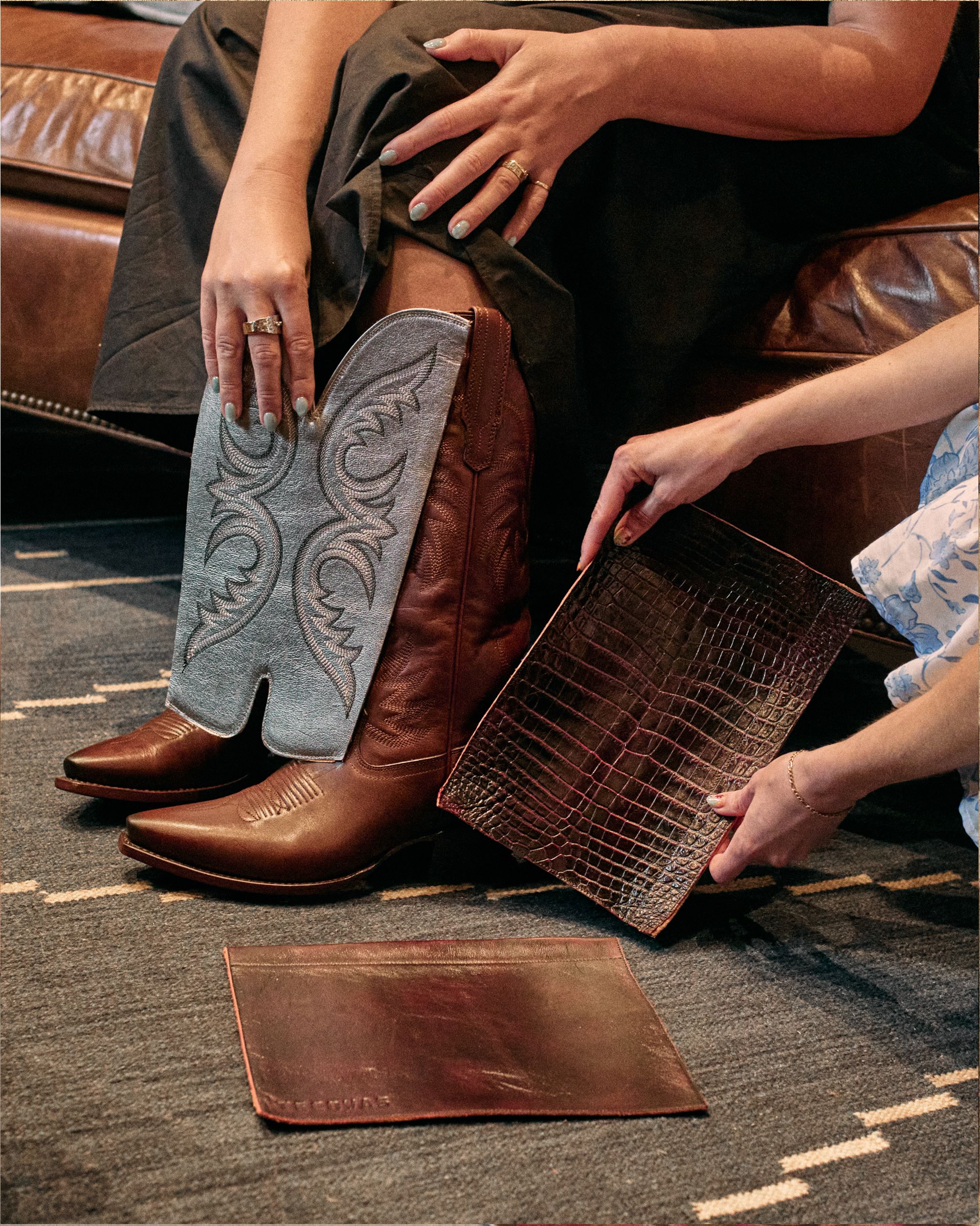 Two people compare leather samples while one wears tall brown cowboy boots with ornate stitching, seated on a leather couch over a patterned rug.