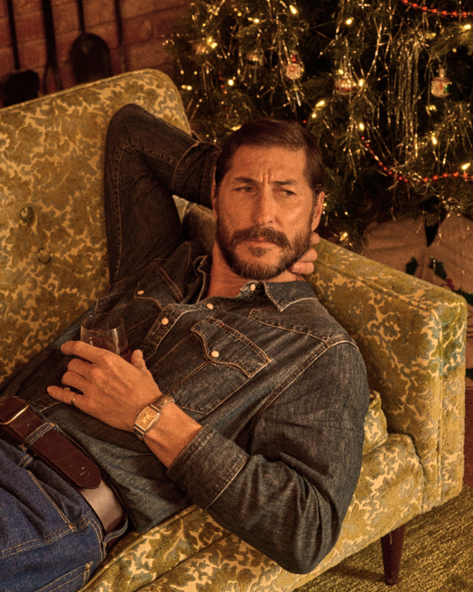 A man in a denim shirt reclines on a patterned green couch, holding a drink, with a decorated Christmas tree in the background.