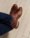 A person wearing brown leather boots and dark blue jeans, seated on a light-colored wooden floor.