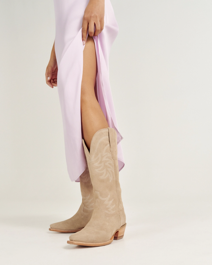 A person in a light purple dress is showing off their beige cowboy boots against a plain background.