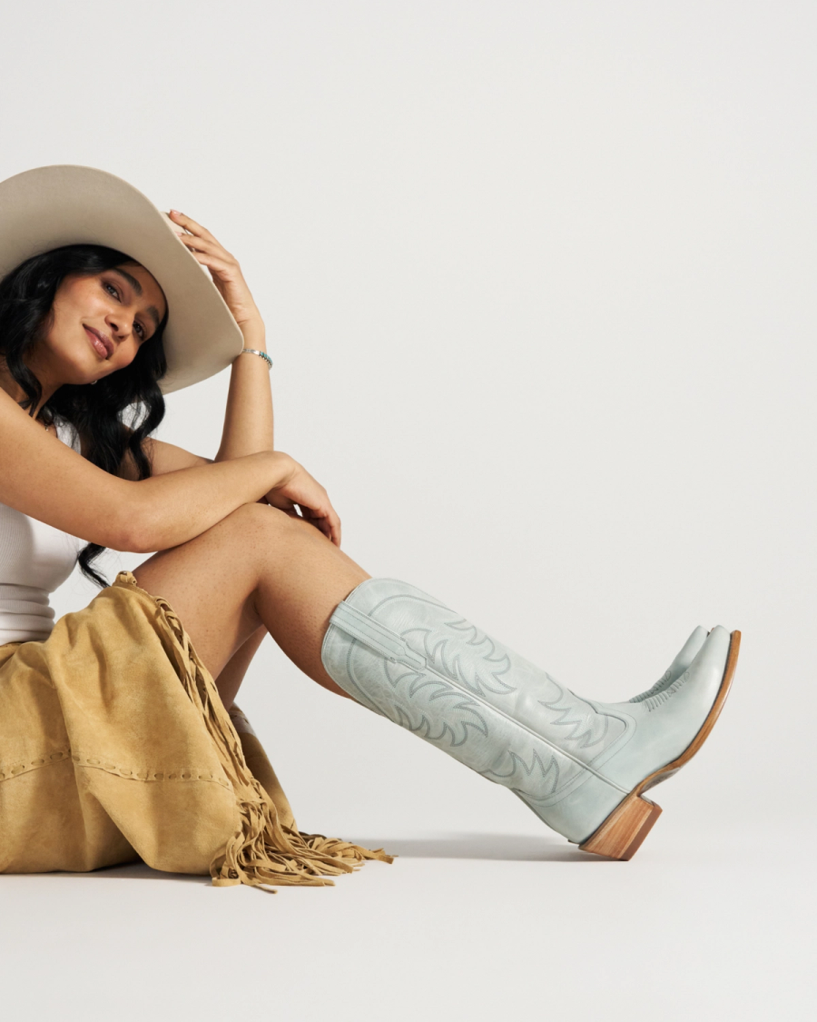 The Annie in Slate Blue Cowhide. Woman in a beige skirt and white tank top sitting on the floor, wearing a white cowboy hat and light blue cowboy boots, against a plain background.