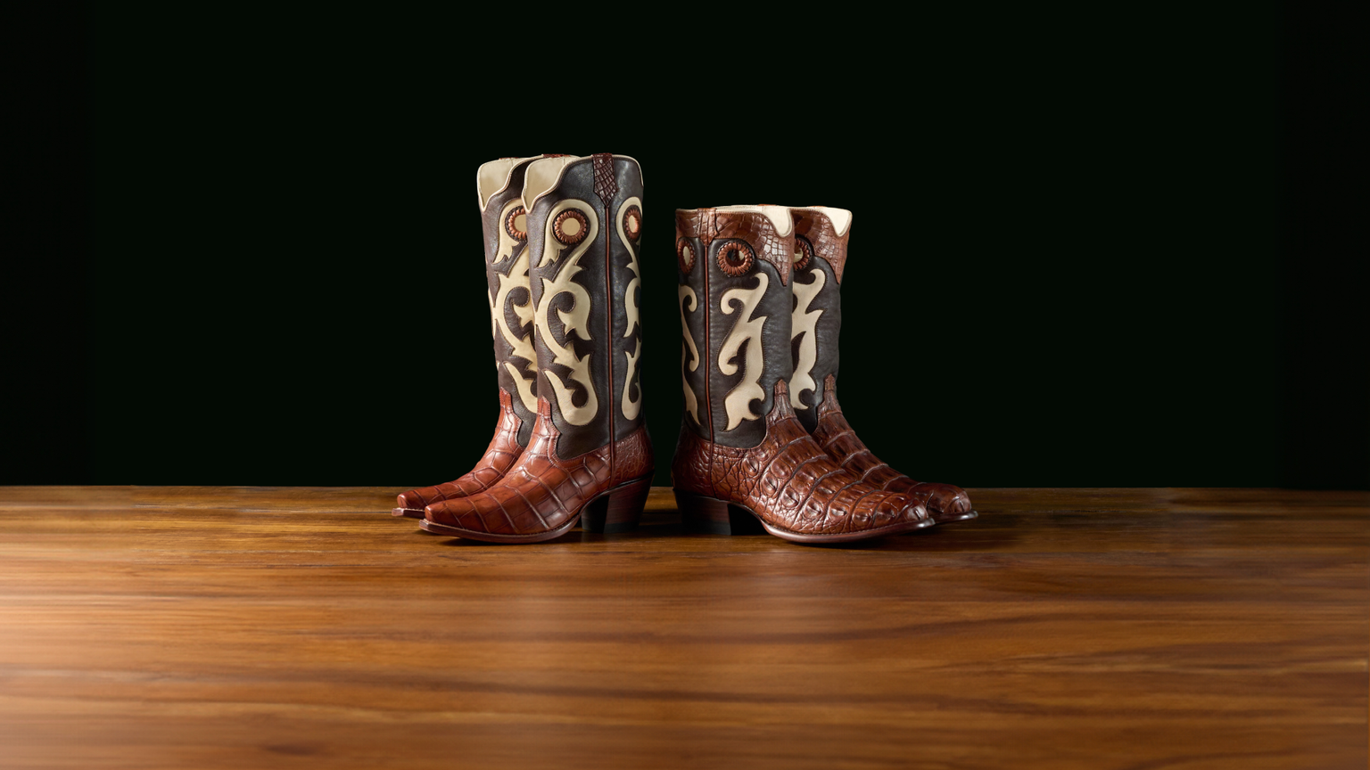 A pair of intricately designed cowboy boots with detailed leather patterns sits on a wooden surface against a dark background.