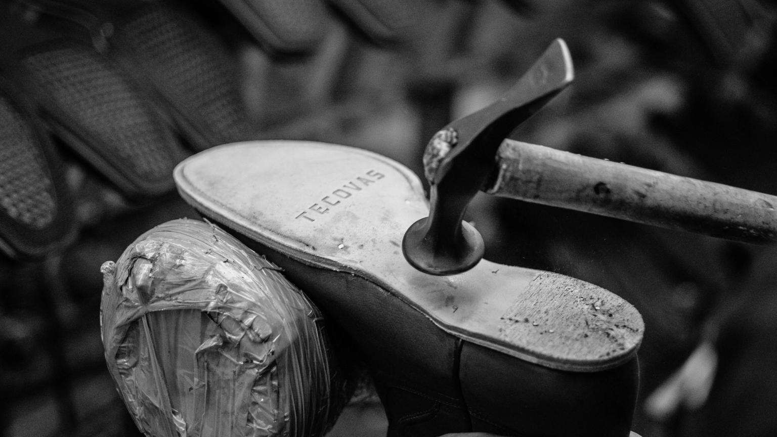 Close-up of a shoe being repaired. A cobbler's tool is positioned at the sole, which is marked "TECOVIS." The shoe's heel is wrapped in protective material. Black and white image.
