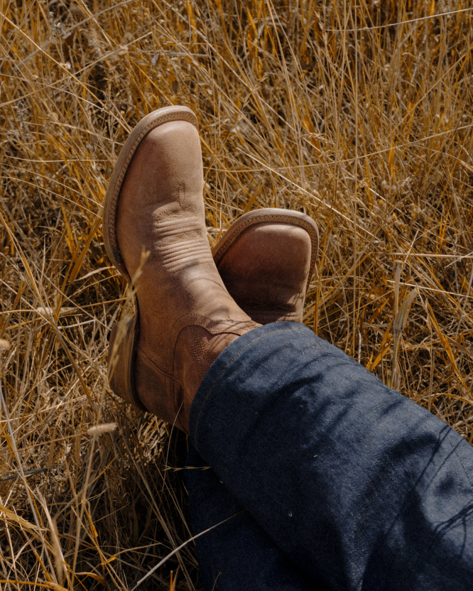 The Doc in Scotch Goat. A person wearing tan leather Scotch Goat square-toe boots and blue jeans, resting their crossed feet in dry grass.