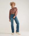 Woman wearing a cowboy hat, long-sleeve shirt, jeans, and boots standing against a plain light background.