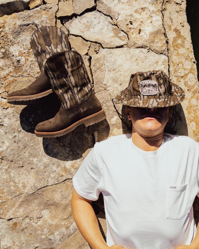 A person in a white T-shirt and camouflage hat lies on a sunlit, cracked stone surface next to a pair of camouflage boots.