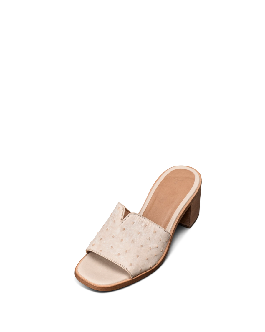 The Carmen Paloma in Pearl Ostrich. A beige open-toe heeled slide sandal, The Carmen Paloma Ostrich, features a wide textured strap and a low wooden block heel, displayed on a plain white background.