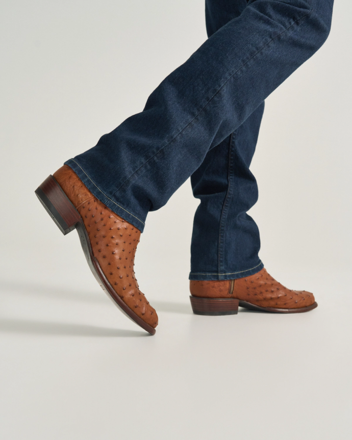 The Dean in Pecan Ostrich. A person wearing blue jeans and distinctive pecan ostrich leather cowboy boots steps forward on a light-colored surface.