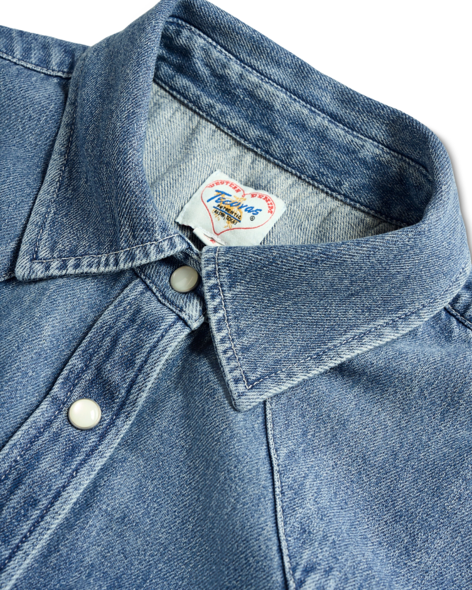 Close-up of a blue denim shirt with a collar, snap buttons, and a Tecovas brand label at the neckline.