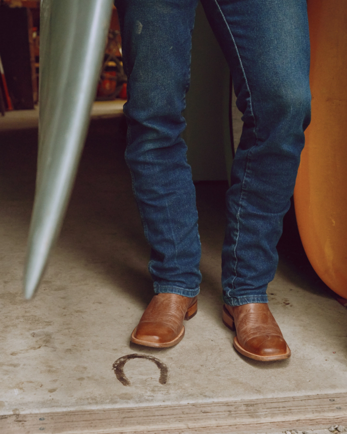 Person wearing blue jeans and brown leather shoes standing on a concrete floor beside an object with a horseshoe on the ground.