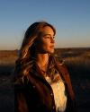 A woman with long hair stands outdoors at sunset, looking into the distance. She is wearing a brown jacket over a white top. Dry grass and open land are visible in the background.