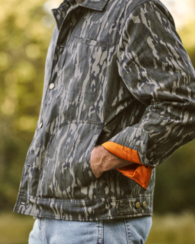 Person wearing a gray camouflage jacket with orange lining and blue jeans, standing outdoors with hands in pockets.