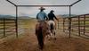 Two people on horseback ride through a metal gate into an open field, with hills visible in the background under a cloudy sky.