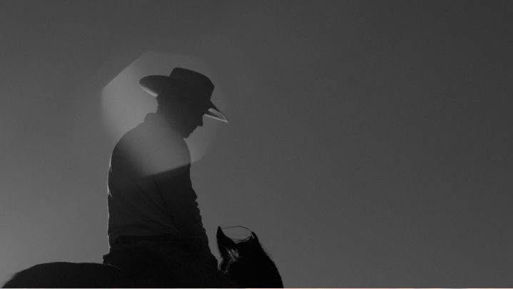 Silhouette of a person wearing a cowboy hat riding a horse against a clear sky with sunlight in the background.