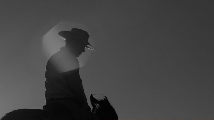 Silhouette of a person wearing a cowboy hat riding a horse against a clear sky with sunlight in the background.