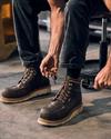 A person ties the laces of brown leather boots while sitting on a stool in a workshop setting.