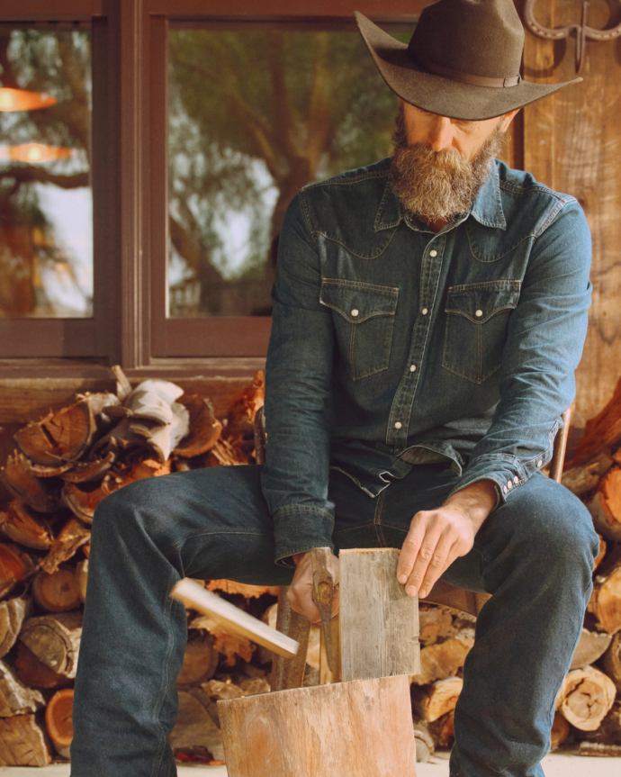 Man in denim outfit and cowboy hat chops wood, seated in front of firewood stack by a window.