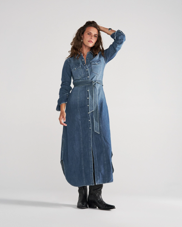 A woman poses in a long-sleeved, belted denim shirt dress with a front button placket and side slits, paired with black boots.