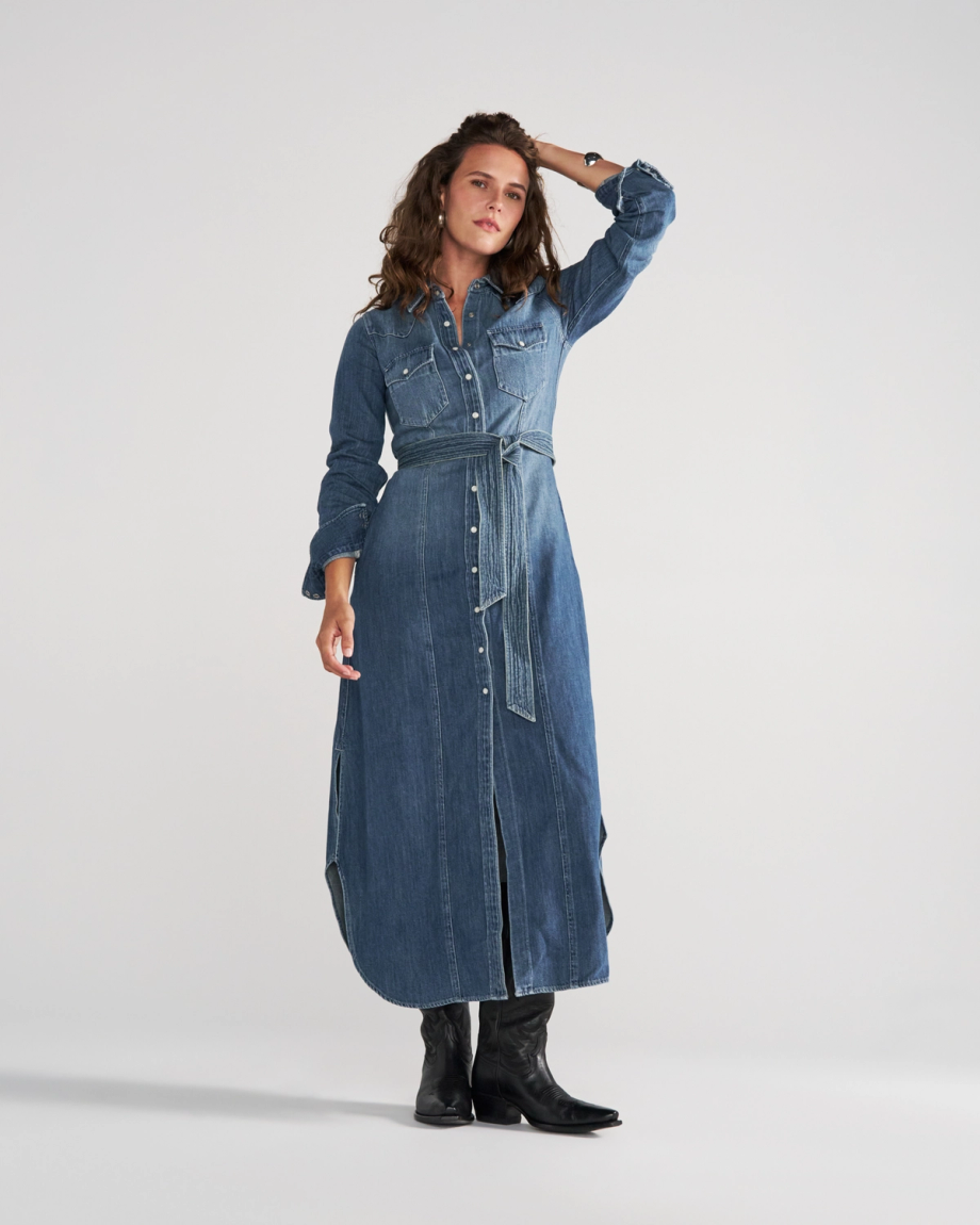A woman poses in a long-sleeved, belted denim shirt dress with a front button placket and side slits, paired with black boots.