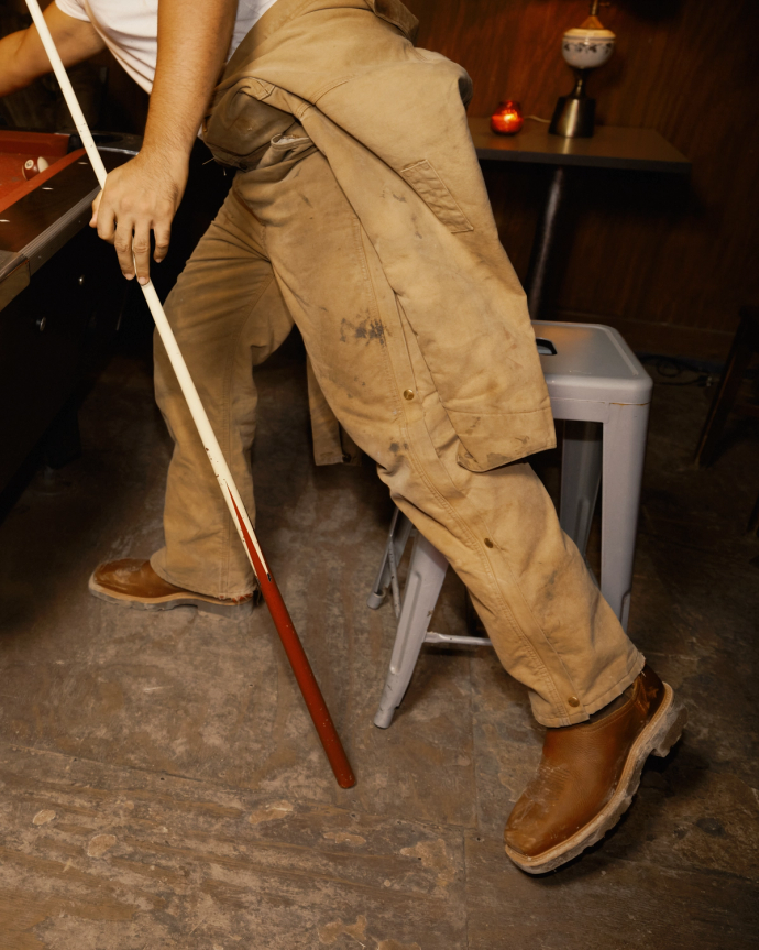 A person in work boots and tan overalls playing pool, holding a cue stick over a pool table.