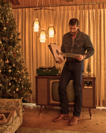 The Cartwright in Scotch Goat. A man in denim clothes examines a vinyl record beside a vintage TV, record player, and decorated Christmas tree in a retro-styled living room, while a glass of Scotch rests on the table next to him.