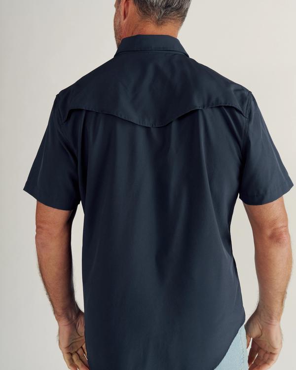 Man wearing a short-sleeve, dark button-up shirt and light blue jeans, shown from the back against a plain background.