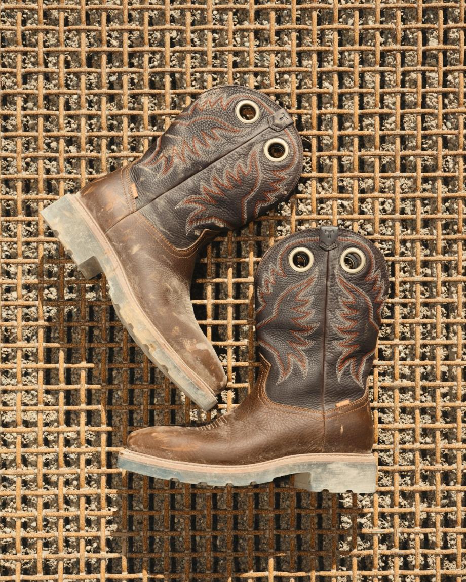 LongHaul Waterproof Square Composite Toe Work Boot in Saddle/Fossil Cowhide. A pair of worn maple cowhide cowboy boots with black and orange stitching rests on a rusty metal grid surface.