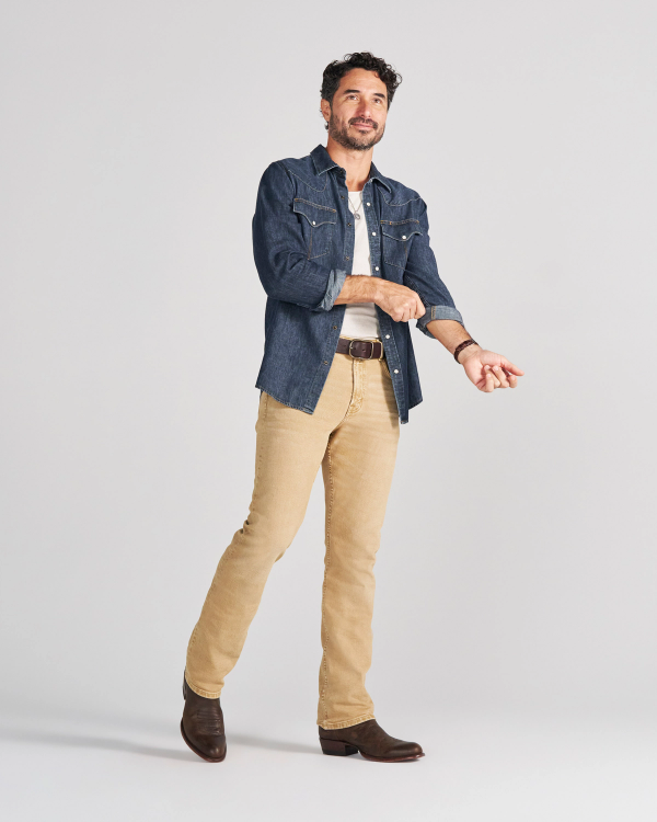 A man in a denim shirt, white t-shirt, tan jeans, and brown boots rolling up his sleeve.