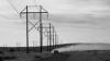 A car drives on a dirt road beneath tall power lines, raising a trail of dust across a flat, grassy landscape under a cloudy sky.