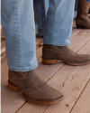 Close-up of a person's legs wearing light blue jeans and brown leather ankle boots, standing on a wooden floor. A second pair of boots is partially visible in the background.