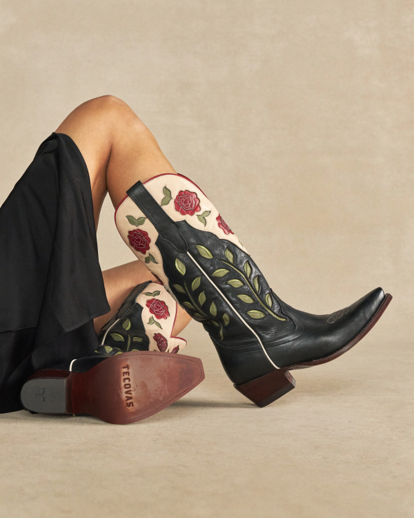 The Annie in Midnight Roses Cowhide. A person in a black skirt reclines on the floor wearing The Annie Cowhide black cowboy boots, adorned with red rose and green leaf embroidery.