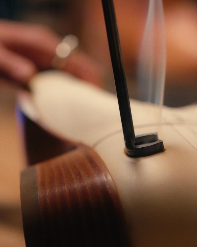 A hot metal tool is being used to burn a straight line onto the sole of a light-colored leather shoe.
