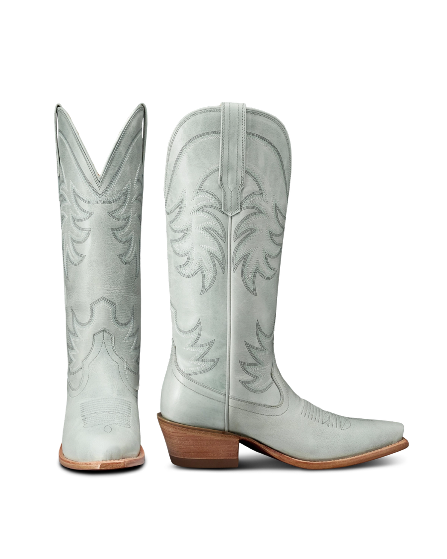 The Annie in Slate Blue Cowhide. A pair of light gray cowboy boots with decorative stitching, pointed toes, and wooden heels; one boot is shown from the back, the other from the side.