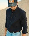 A man wearing a dark button-up shirt, light blue jeans, and a dark cap with a patch, standing outdoors on gravel, looking down.