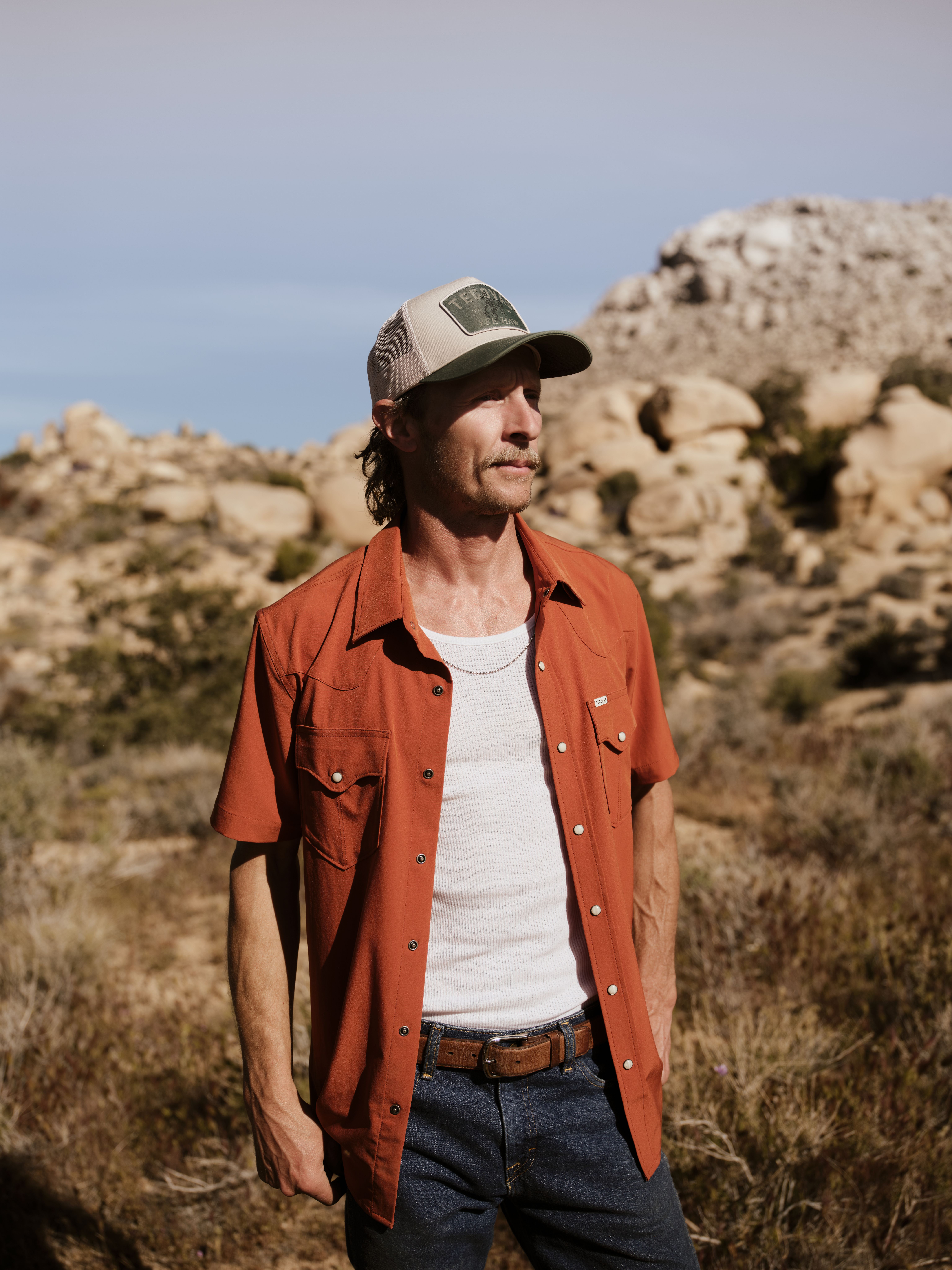 Man wearing a rust-colored short-sleeve shirt, white tank top, and cap stands outdoors in a rocky desert landscape.