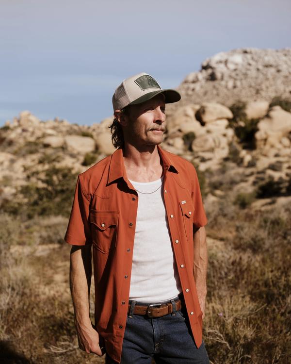 Man wearing a rust-colored short-sleeve shirt, white tank top, and cap stands outdoors in a rocky desert landscape.