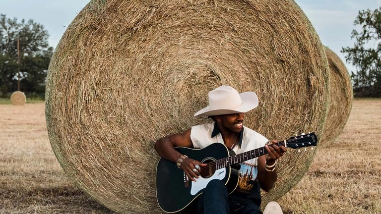 Image of man playing guitar in a field leaning against a barrel of hay.