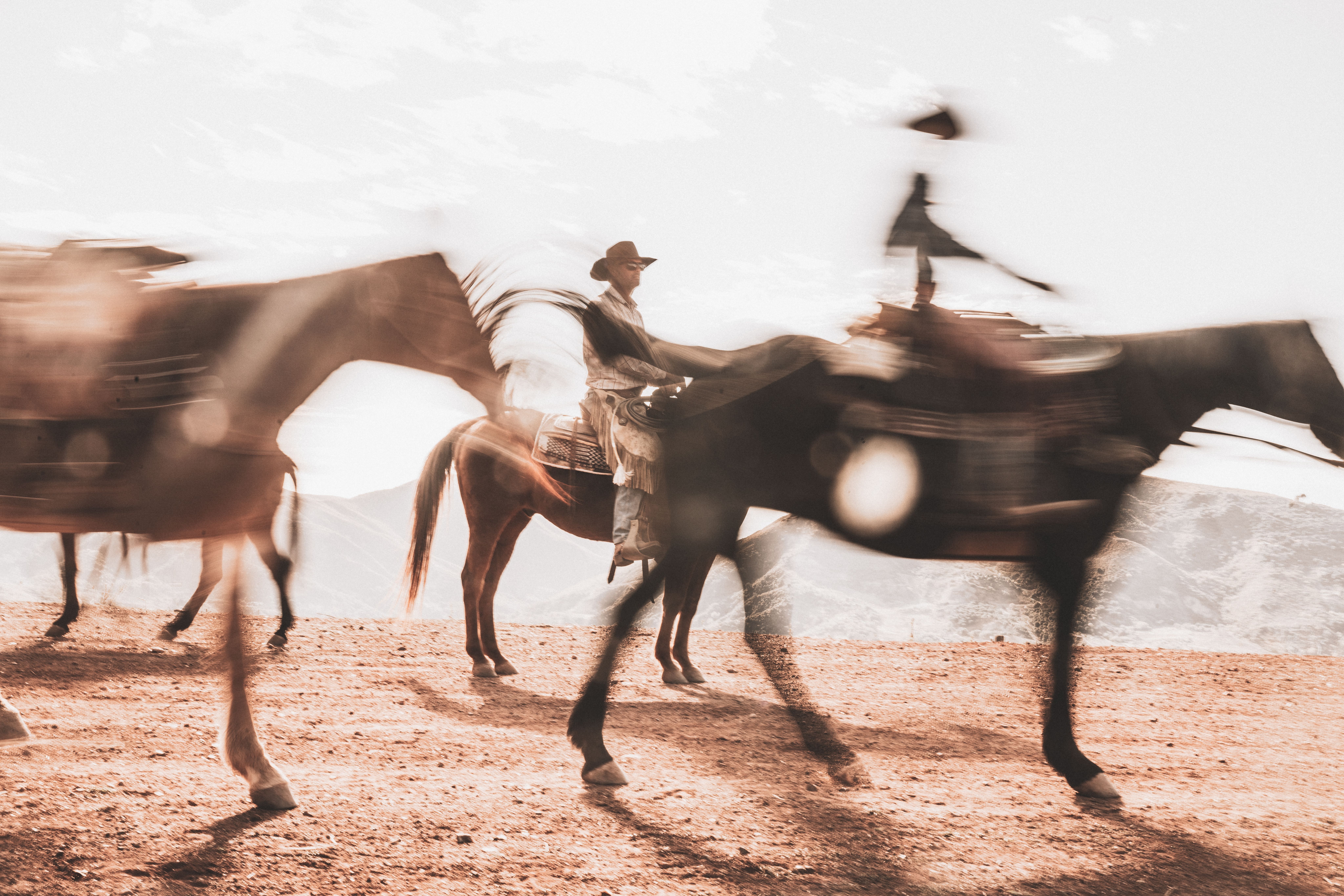 Three people ride horses across a dry, dusty landscape with motion blur, under bright daylight and distant mountains.