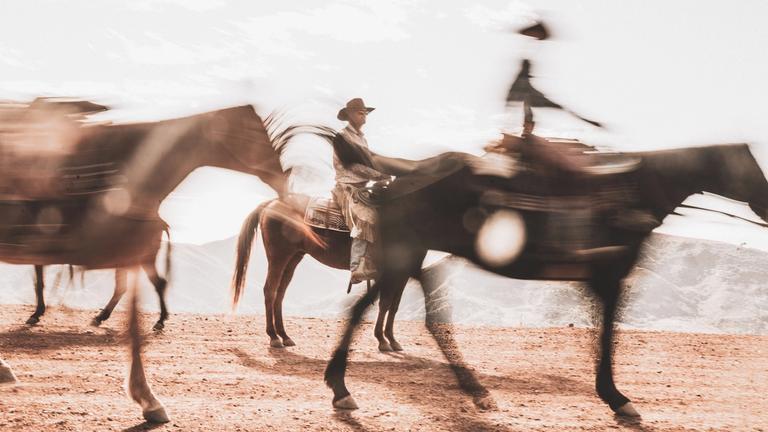 Three people ride horses across a dry, dusty landscape with motion blur, under bright daylight and distant mountains.