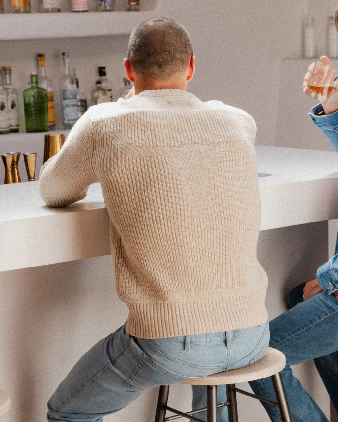 A person in a beige sweater and blue jeans sits on a stool at a white bar counter, holding a drink.