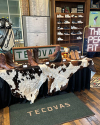 Display of Tecovas boots on cowhide rugs with a variety of styles on a table and a shelf in the background. Golf bags and a neon sign are also visible.