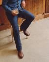 A person in blue jeans and a denim shirt sits on a wooden stool, holding a glass of amber liquid, with brown leather boots on a concrete floor.