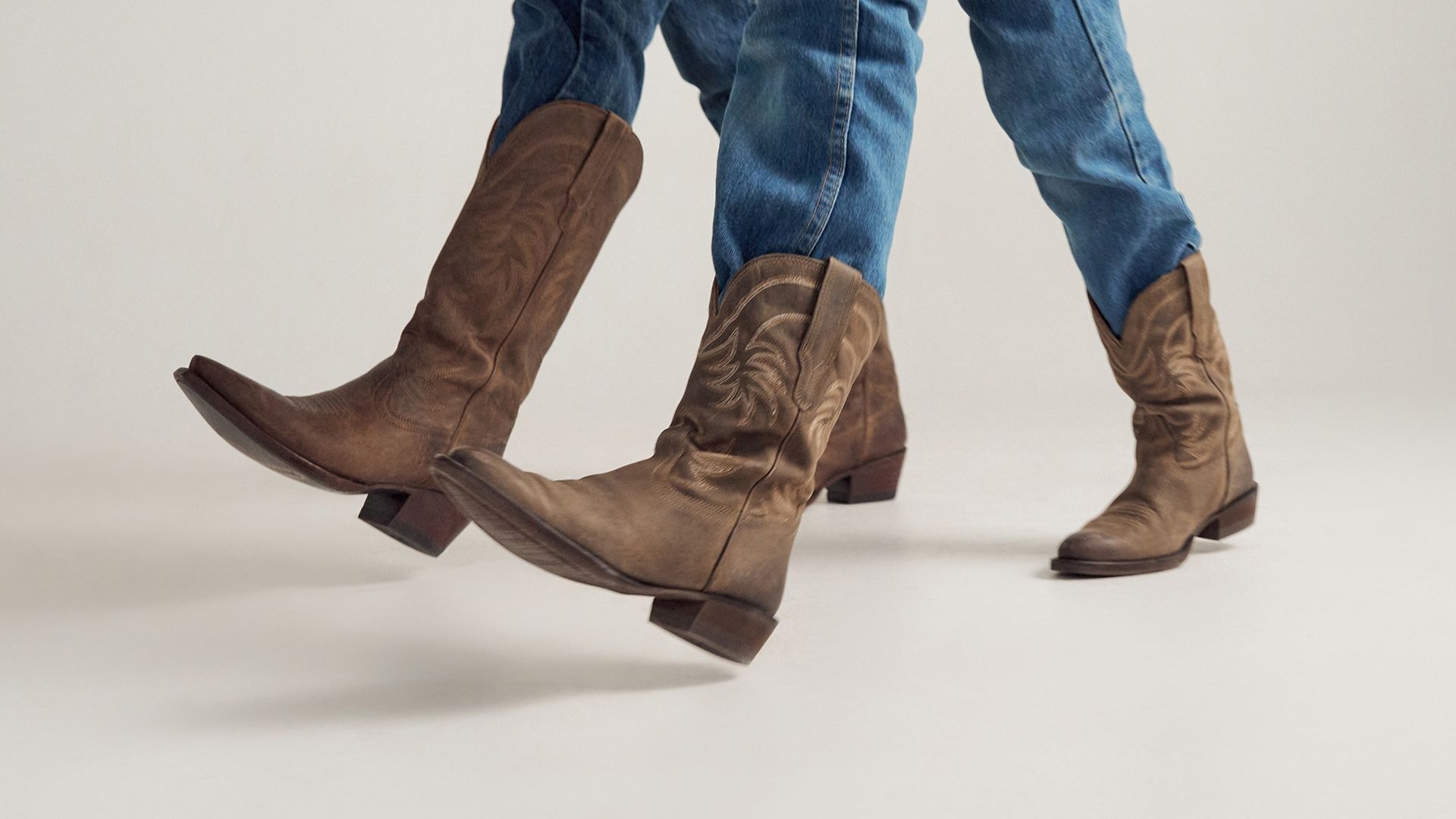 Two people wearing blue jeans and brown cowboy boots walk forward on a plain light background.