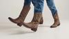 Two people wearing blue jeans and brown cowboy boots walk forward on a plain light background.