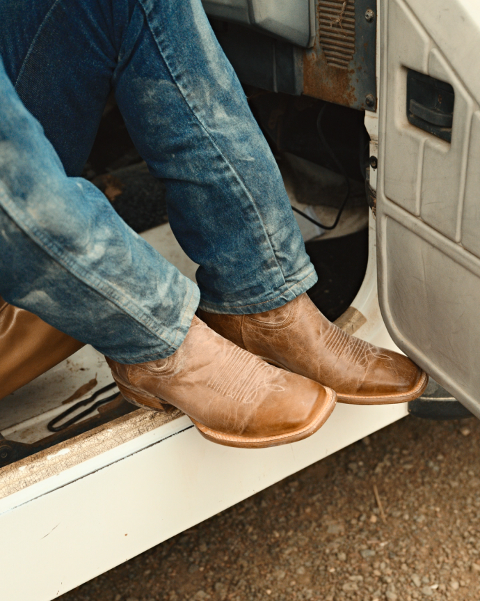 Person wearing dusty brown boots and blue jeans, seated in a vehicle with feet on the door frame.